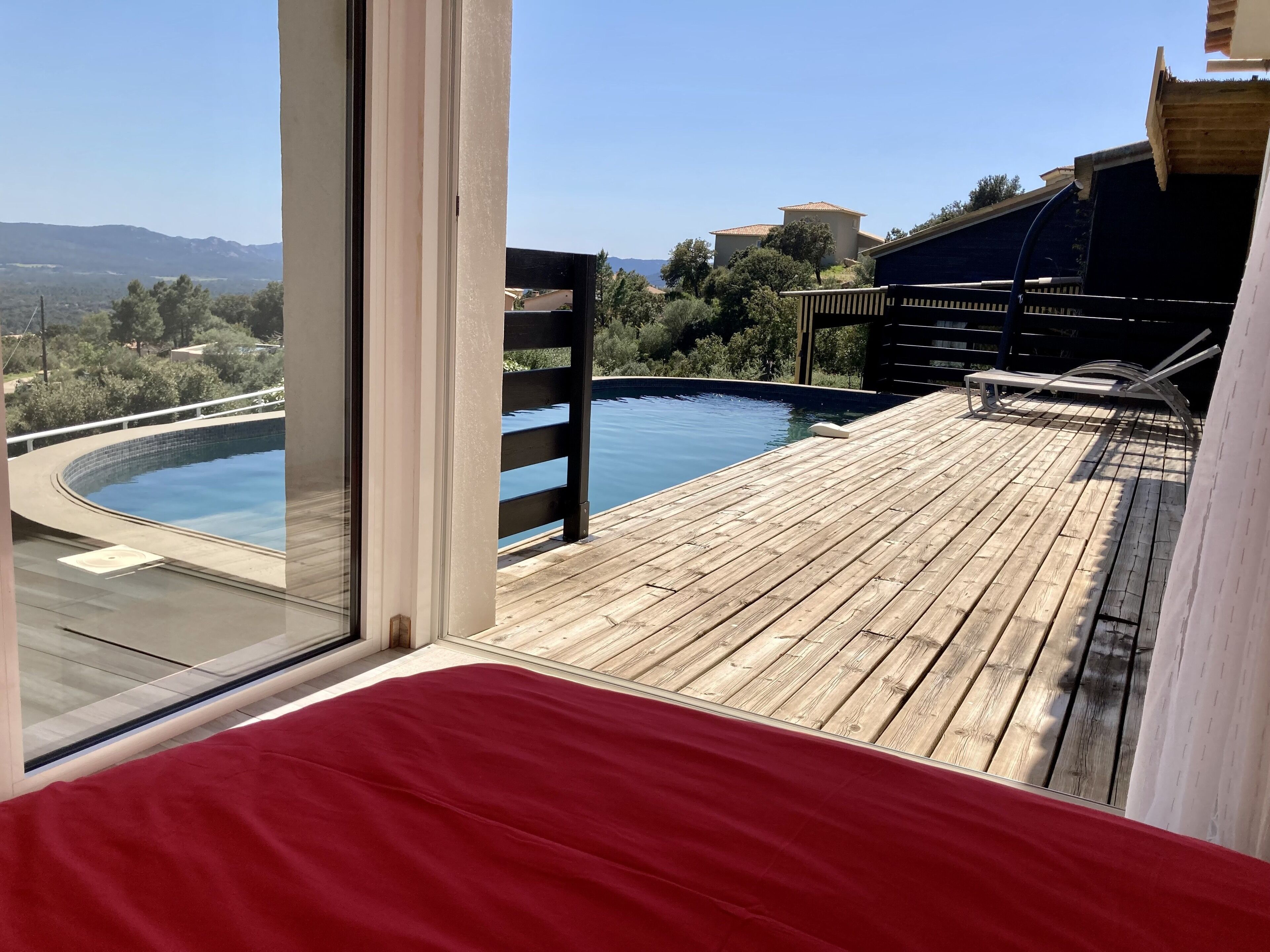 A wooden deck with a pool, a red bed, and a view of the landscape.