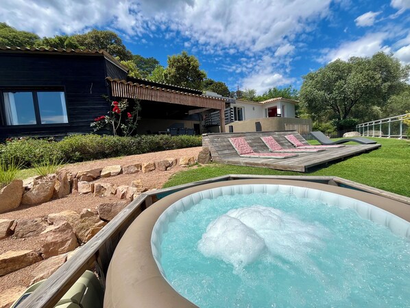 A backyard with a hot tub, a wooden deck, and a house with a sloped roof.