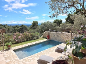 A stone pool with lounge chairs, surrounded by a stone wall and potted plants, overlooking a scenic landscape with trees and mountains.