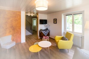 Ein Wohnzimmer mit einem gelben Sessel, einem gemusterten Sessel und einem kleinen runden Tisch. Im Hintergrund ist eine Treppe zu sehen.