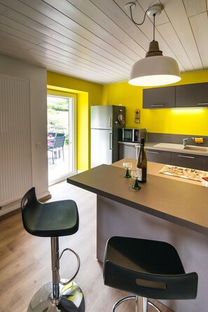 Eine moderne Küche mit gelber Akzentwand, einer zentralen Kochinsel mit Barhocker, einer Hängeleuchte und Blick ins Freie.