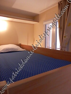 Una cama individual con colchón azul y cabecera de madera en una habitación con paredes color beige y una ventana con cortinas.