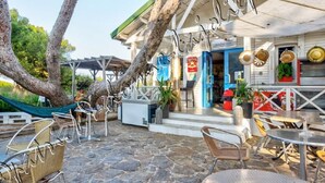 An outdoor seating area with stone flooring, metal chairs, and tables. There are trees and a hammock in the background. - Hyères on the Peninsula Giens, Beach and swimming pool facing Porquerolles *** ! (Hyères)