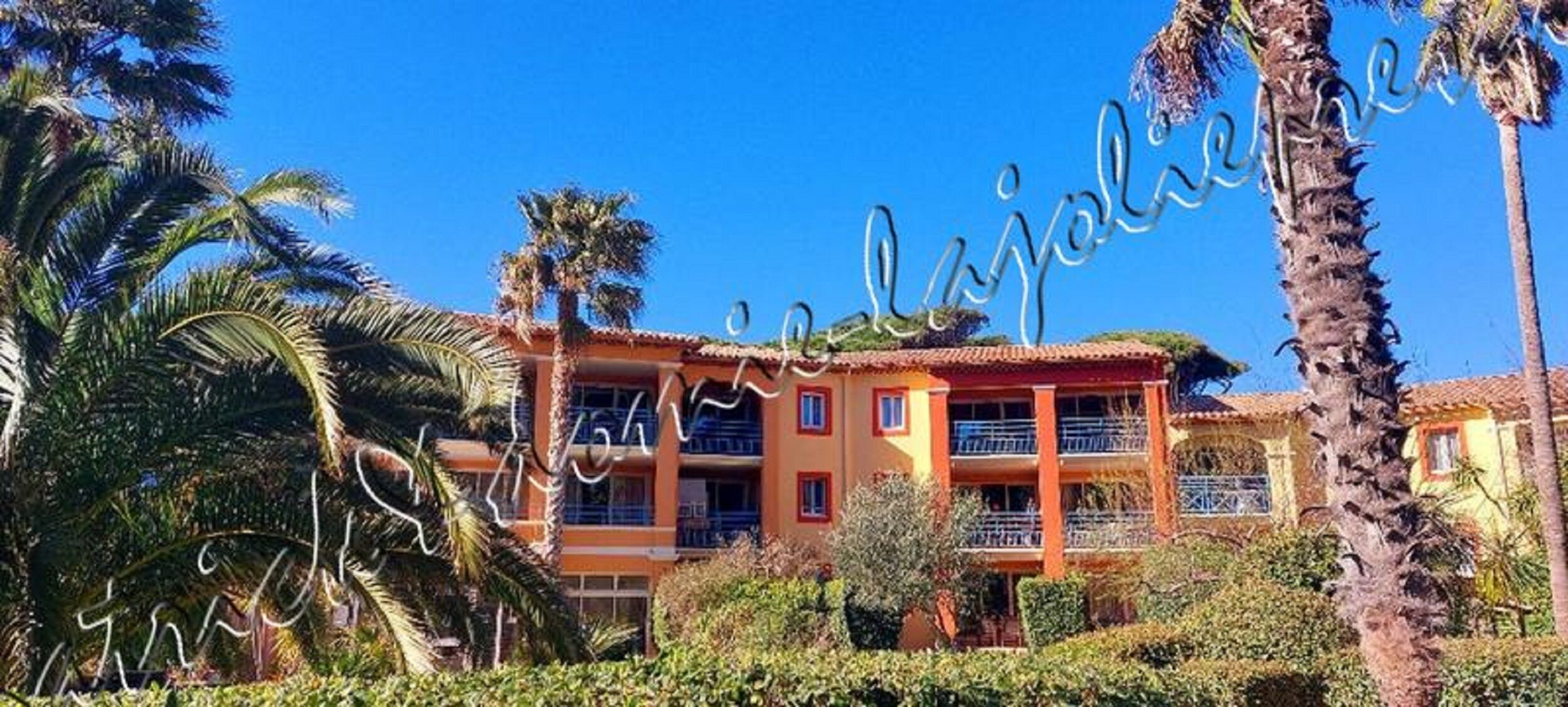 A multi-story hotel with balconies, surrounded by palm trees and greenery.