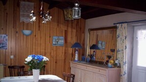 A dining area with a wooden wall, a table with a white tablecloth, chairs, a cabinet with decorative items, and a mirror with a floral curtain. - house by the sea on the Vendée coast quiet good plan (Bouin)