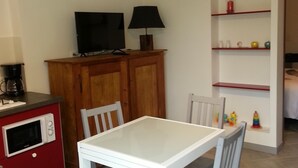 Un pequeño comedor con una mesa y sillas blancas, un mueble de madera con televisor y lámpara, y una estantería con juguetes.