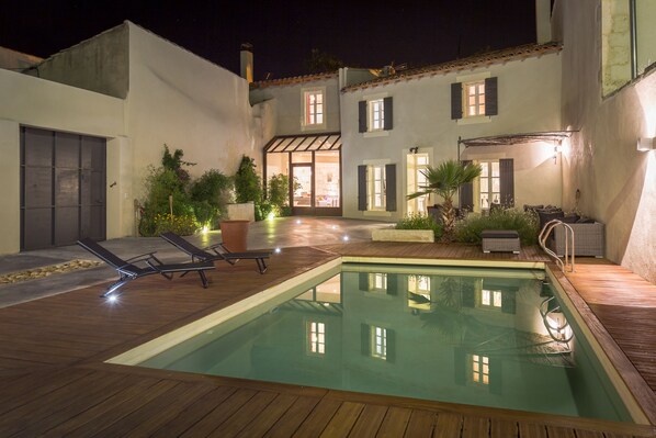 Une piscine avec des chaises longues, une terrasse en bois et un bâtiment doté de nombreuses fenêtres et portes.