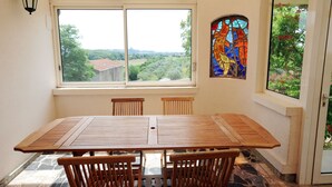 A dining area with a large table and chairs, a stained glass window, and a view of greenery and a house. - Hameau-Montplaisir, Béziers (Béziers)