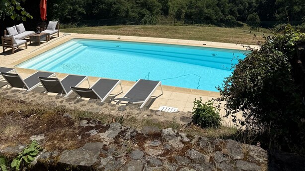 Ein rechteckiger Swimmingpool mit klarem, blauem Wasser, umgeben von einer Steindeck. In der Nähe des Pools befinden sich Liegestühle und ein roter Sonnenschirm. Im Hintergrund erheben sich üppige, grüne Hügel und ein klarer, blauer Himmel.