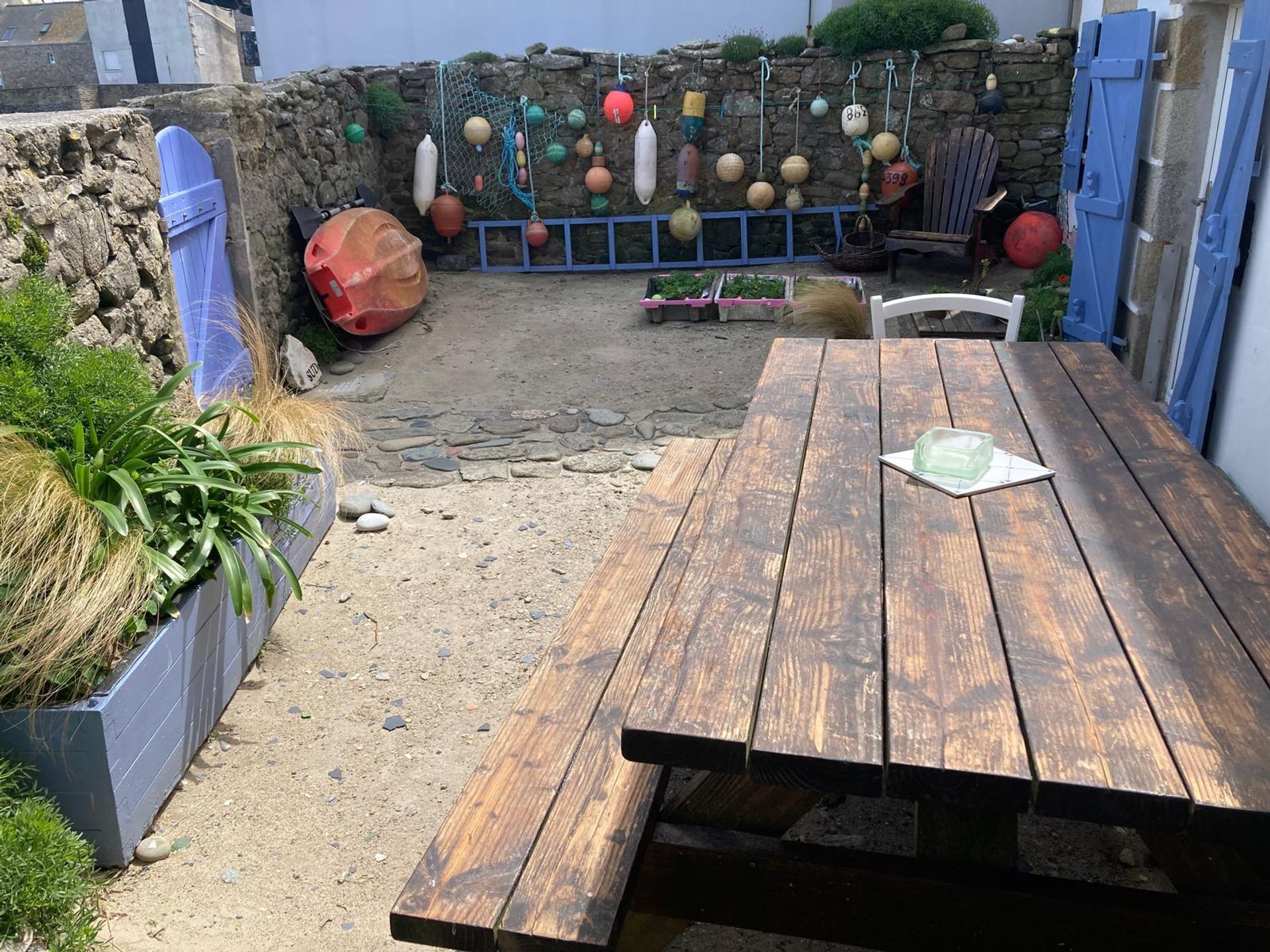 Une table en bois, dans un décor extérieur rustique, avec un mur de pierres et divers bouées colorées suspendues au mur.