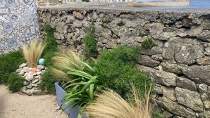 Un muro costiero in pietra con un mosaico in piastrelle, una zona erbosa con piante e vista su edifici e sul mare sullo sfondo.