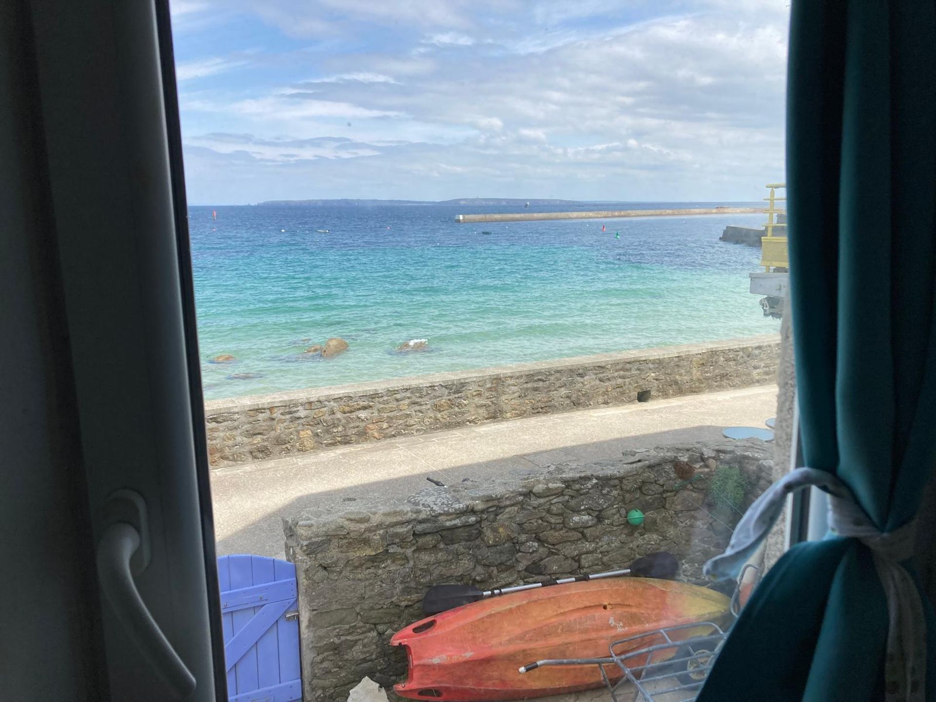 Une vue d’une plage avec une eau turquoise et limpide, un mur de pierre et une porte bleue.
