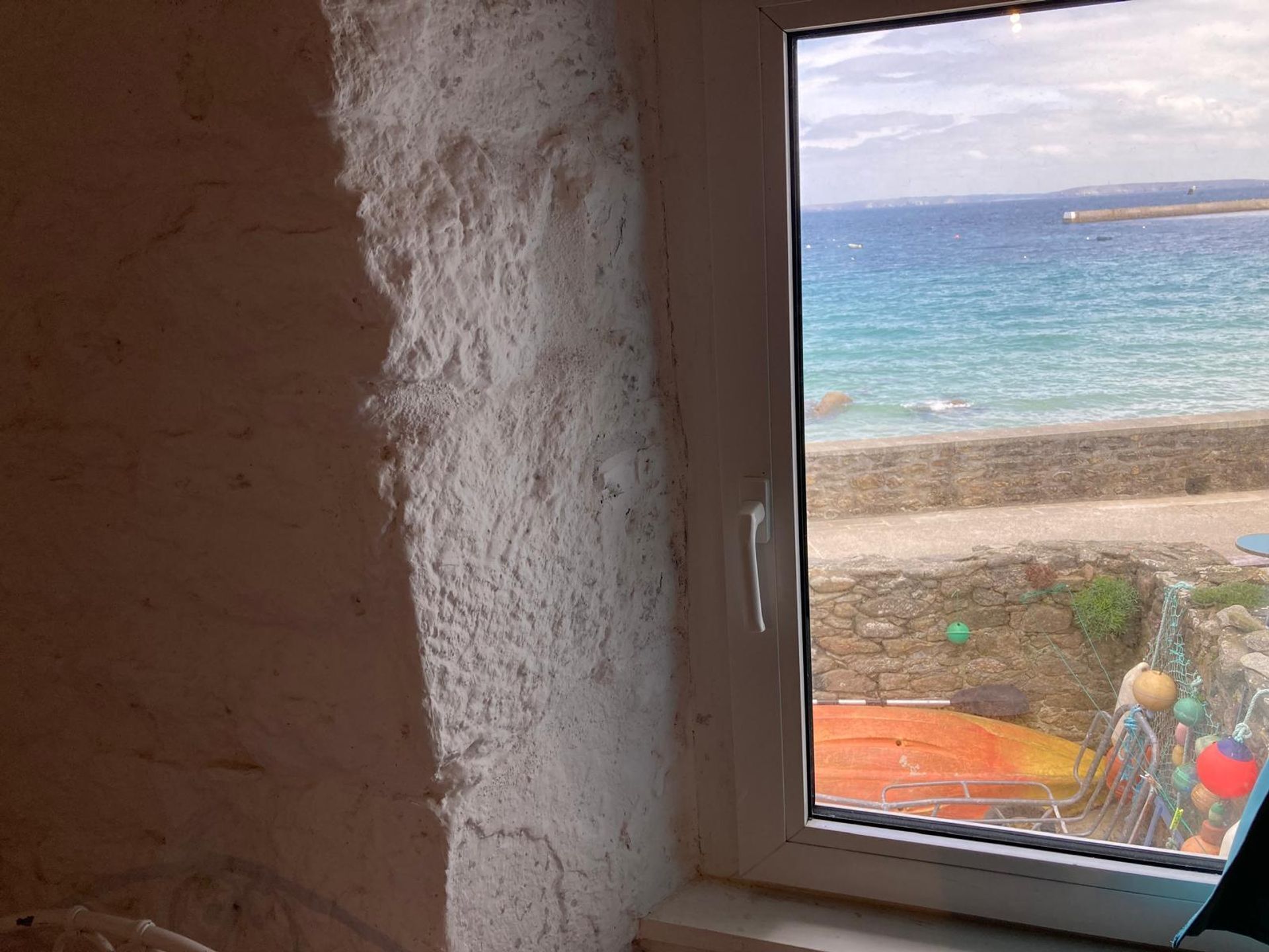 A room with a view of the sea through a window, a stone wall, and a small outdoor area with colorful decorations.