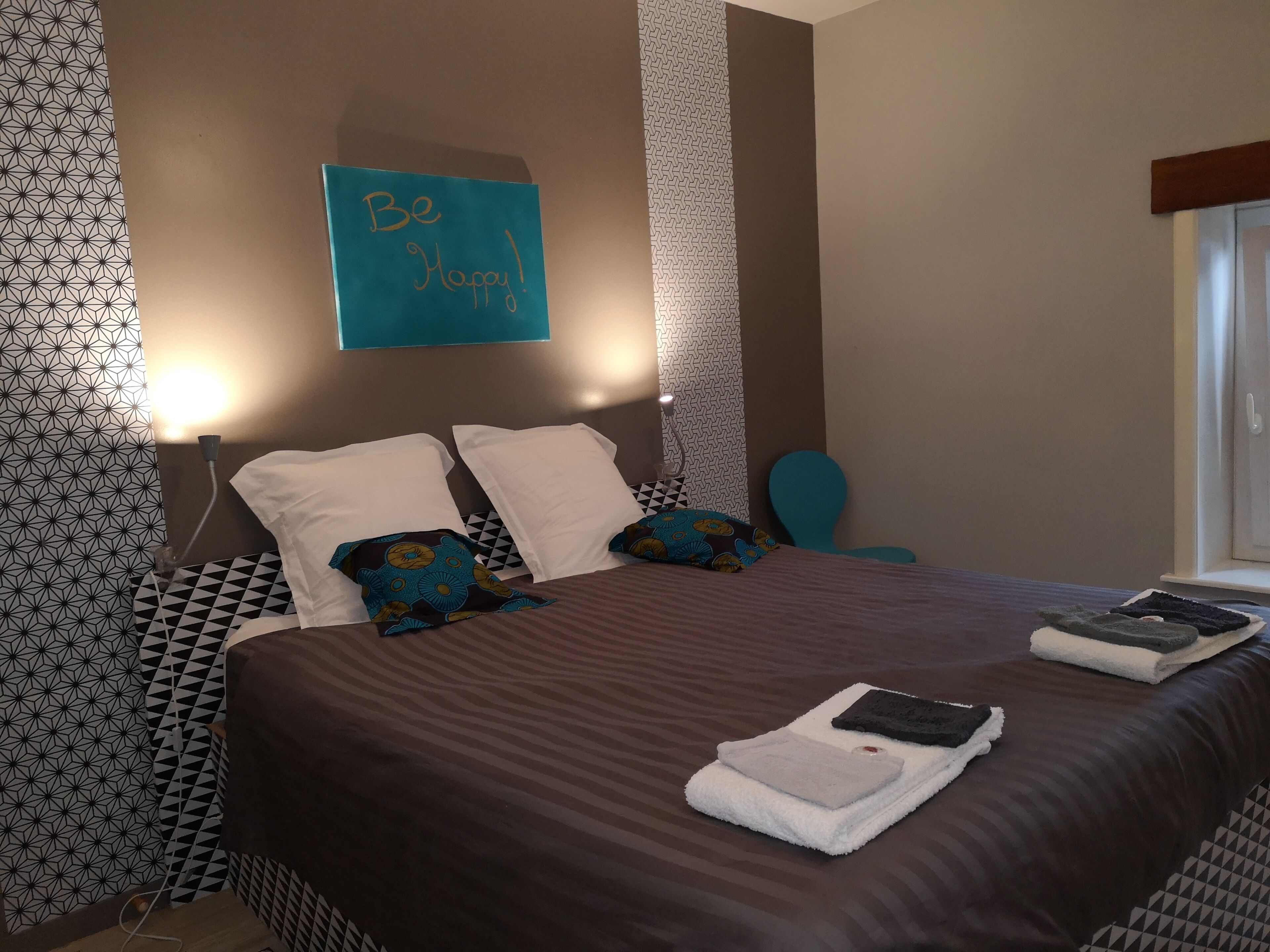 A neatly made bed with a brown bedspread, two white pillows, and decorative pillows with blue and yellow patterns. A sign above the bed reads "Be Happy!" The room has patterned wallpaper and a blue chair.