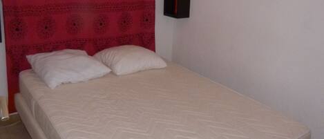 Une chambre avec un lit simple, une tête de lit rouge à motifs, du linge de lit blanc et une petite table de chevet noire sur laquelle se trouve un objet rouge.
