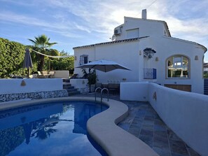A swimming pool with a curved edge, a white building with a tiled roof, and a palm tree in the background. - Character villa for families in residential area with private pool. (JAVEA)
