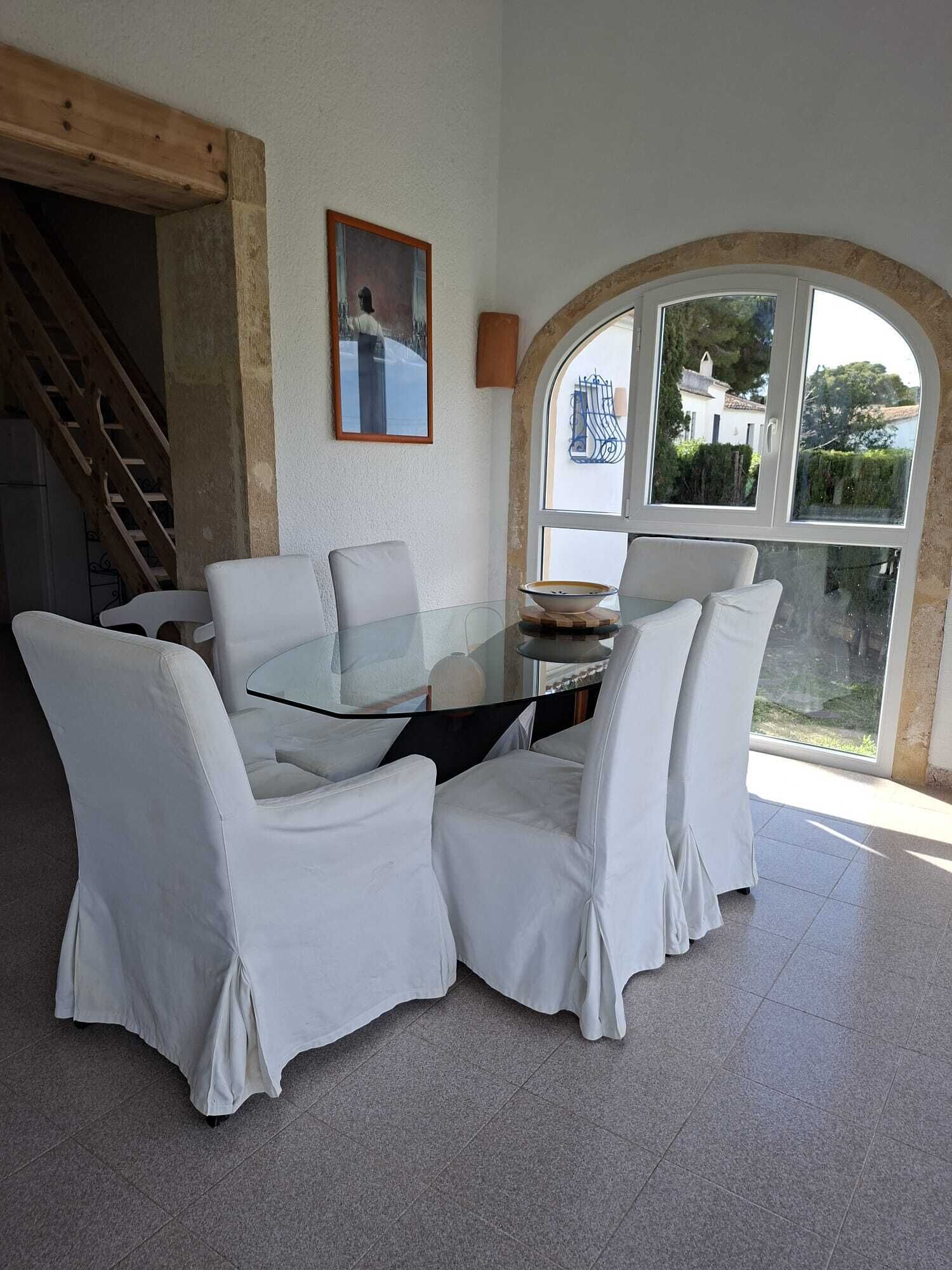 A dining area with a glass table surrounded by white chairs, a painting on the wall, and a view through a large arched window.