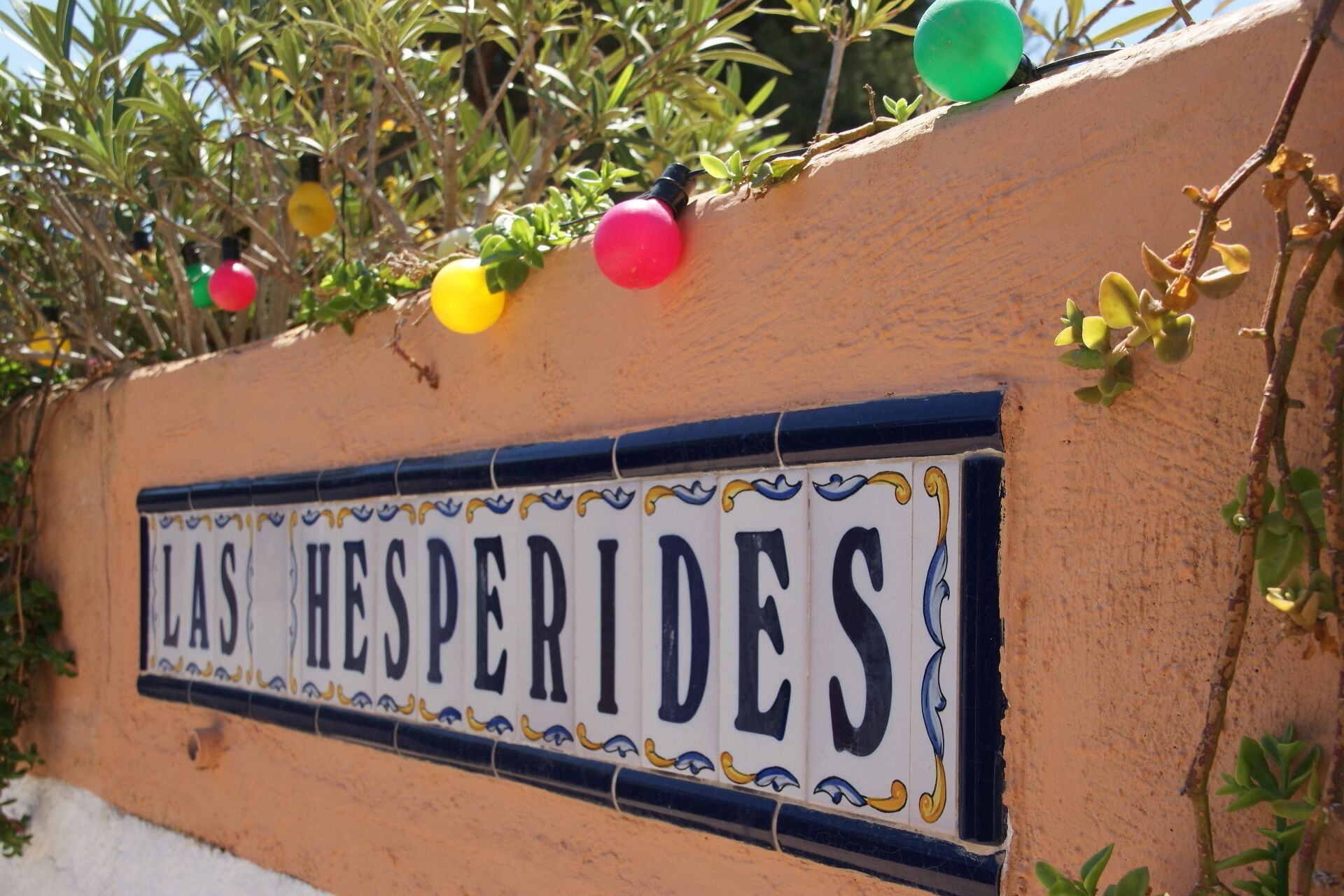 Ein Schild mit dem Schriftzug „LAS HESPERIDES“ an einer Wand, umgeben von Grünpflanzen und Lichterketten.