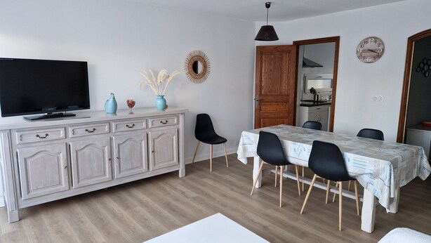 Un comedor con una mesa dispuesta para cuatro, un televisor sobre un mueble, y una cocina al fondo.