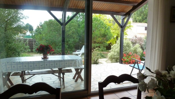 Un patio couvert avec une table et des chaises, une vue sur un jardin agrémenté d’arbres et de fleurs, et un sol carrelé.
