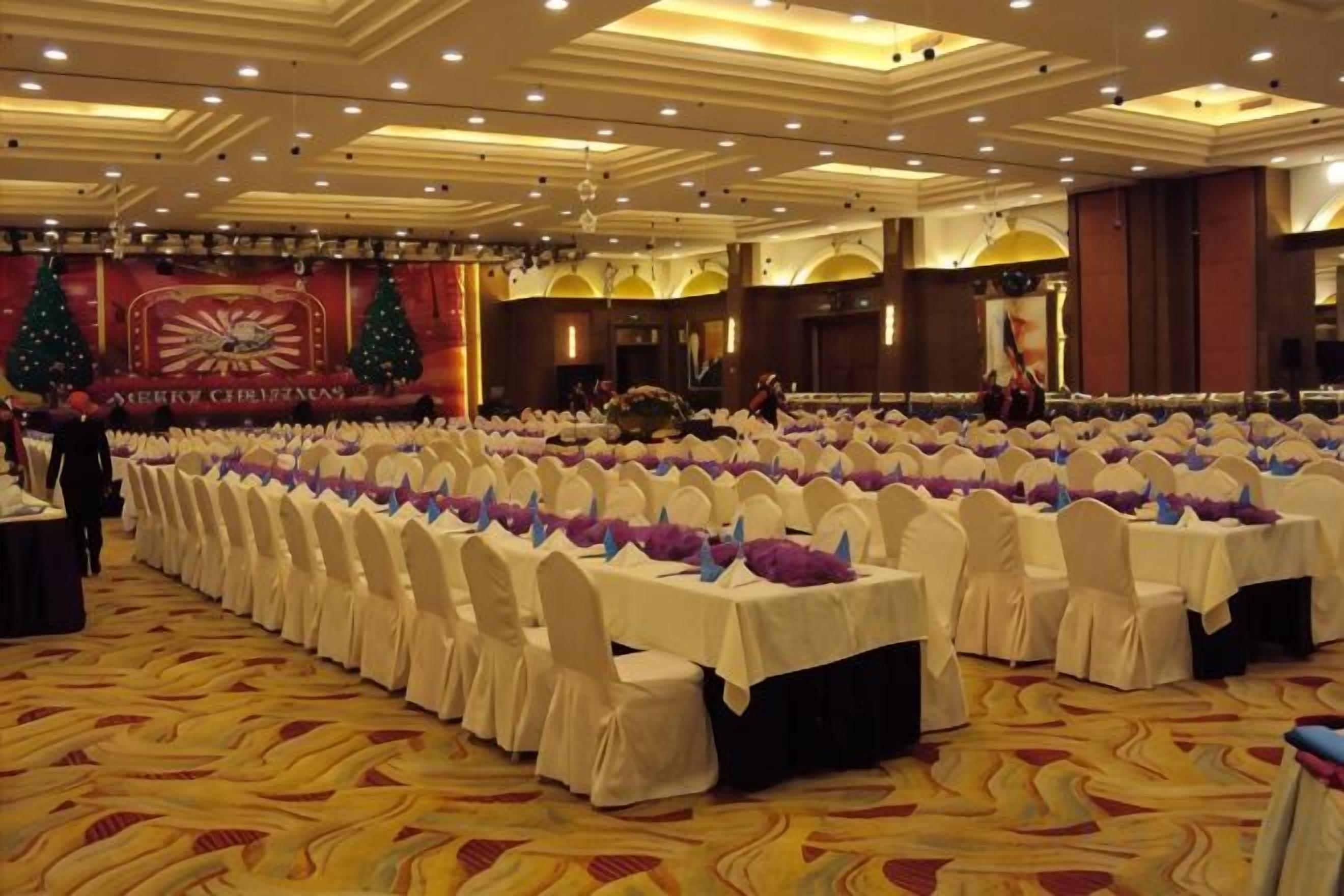 Un salón de banquetes con filas de mesas dispuestas para un evento formal, con piso alfombrado y un escenario decorado de forma festiva.