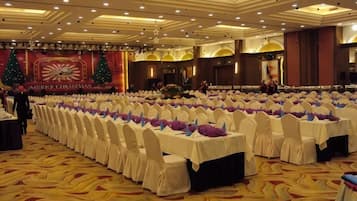 Un salón de banquetes con filas de mesas dispuestas para un evento formal, con piso alfombrado y un escenario decorado de forma festiva.