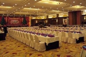 Un salón de banquetes con filas de mesas dispuestas para un evento formal, con piso alfombrado y un escenario decorado de forma festiva.