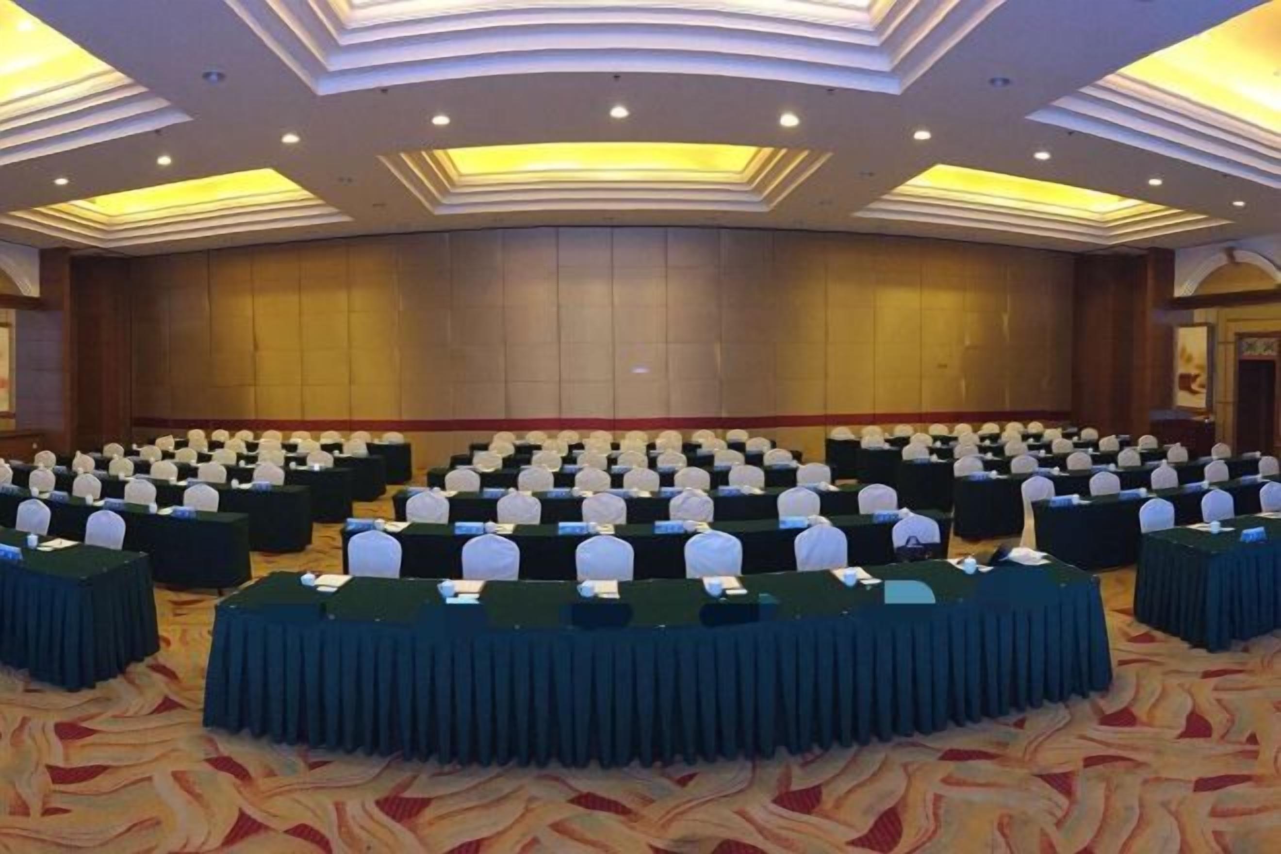 A large conference room with rows of tables and chairs, a stage, and a high ceiling with recessed lighting.