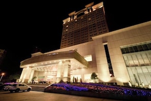 A hotel with a large entrance, a parking area, and a building in the background. - Perfect International Hotel (Qidong)