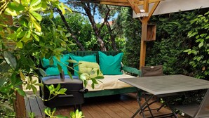 Un patio exterior con terraza de madera, zona de estar cubierta con cojines verdes y una mesa con sillas.
