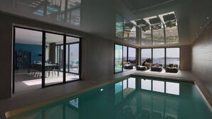 Una piscina interior moderna con paredes de vidrio, un comedor con mesa y sillas, y un área de descanso con sofás.