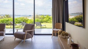 Una sala de estar moderna con un amplio ventanal corredera que da a un balcón con vistas al mar y al follaje. Hay dos sillones y una mesita.