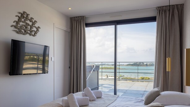 Habitación de hotel con cama, televisión, balcón con vista al mar y un amplio ventanal.