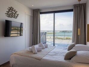 A hotel room with a bed, a TV, a balcony with a view of the sea, and a large window.