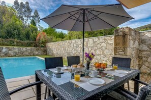 A dining table with a glass top, surrounded by wicker chairs, under a large umbrella.