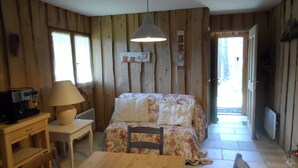 A cozy room with wooden walls, a bed with floral bedding, a table, a chair, a lamp, and a small shelf with decorative items.