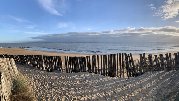 Una playa arenosa con una cerca de madera en primer plano y un mar tranquilo al fondo.