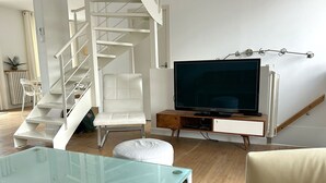 Een moderne woonkamer met een glazen salontafel, een witte bank, een witte fauteuil, een televisie op een houten dressoir en een spiltrap.
