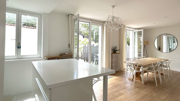 Een lichte keuken met een groot eiland, een eethoek met een houten tafel en stoelen, en grote ramen die voor natuurlijk licht zorgen.