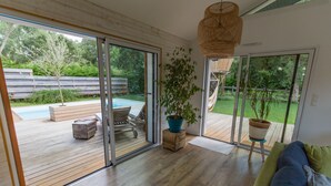A modern living room with a wooden floor, a green sofa, a large potted plant, and a view of a backyard with a pool and outdoor furniture. - Villa 6 pers / with heated pool / near golf & beach / Holiday home (Moliets-Et-Maa)