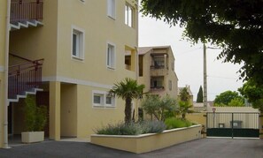 A multi-story residential building with a yellow facade, a gated entrance, and a small garden area with plants.