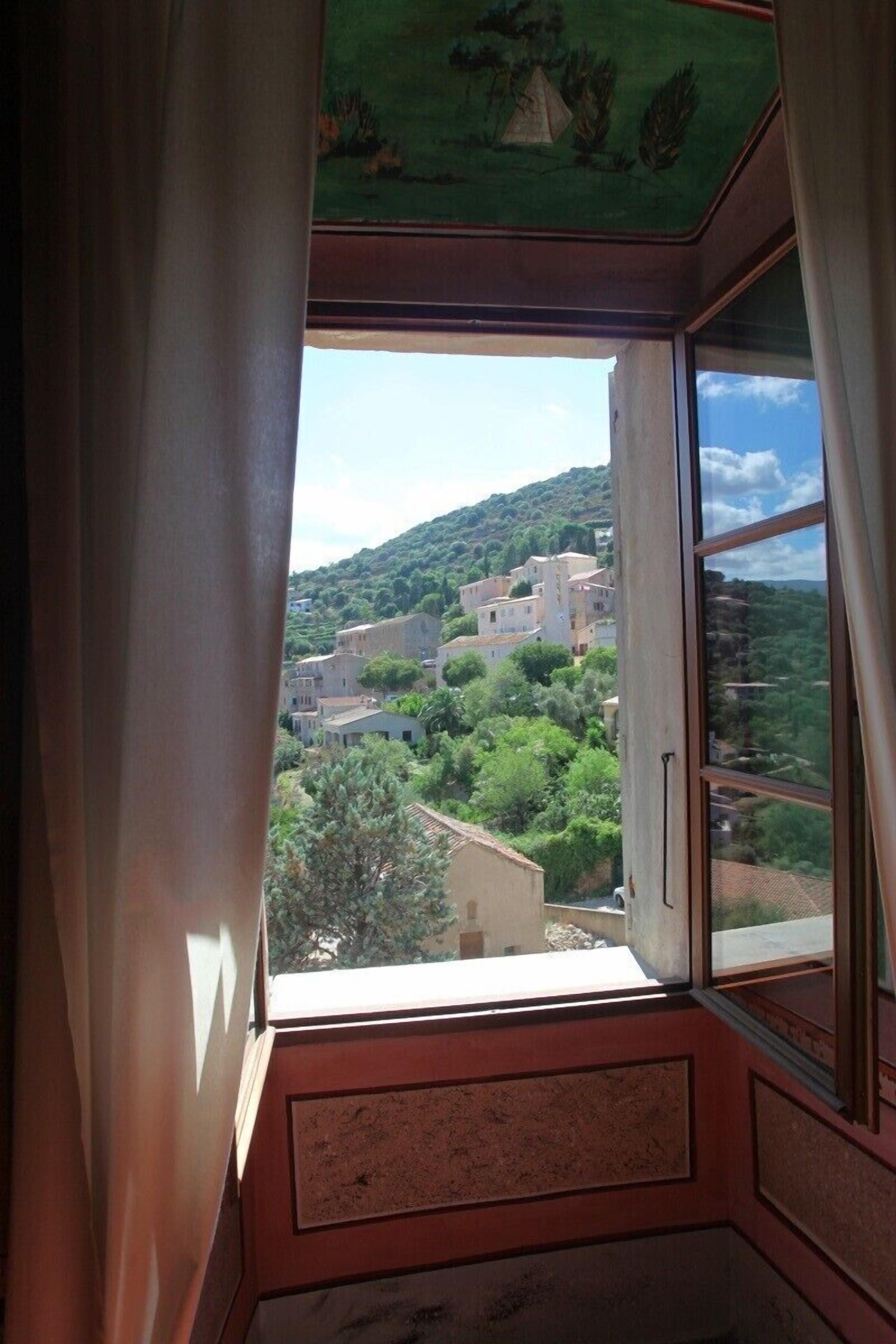 Una habitación con vistas a un pueblo en la ladera, visible a través de una ventana con cortinas.