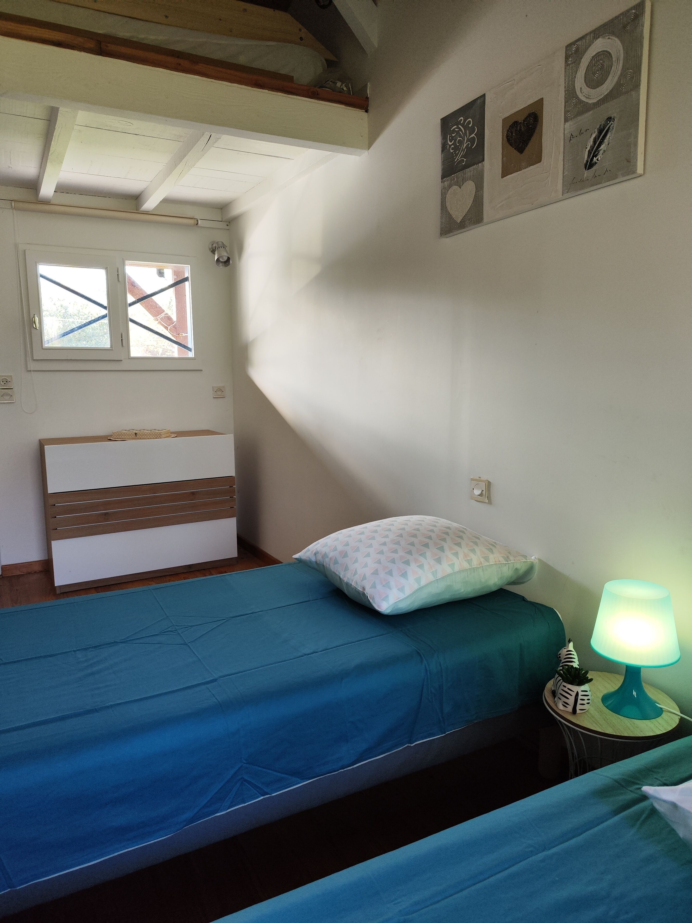 Une chambre à coucher avec un lit, une table de chevet avec une lampe, une fenêtre et des œuvres d’art murales.