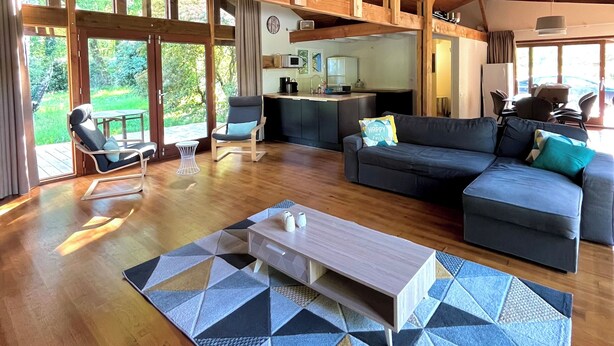 Un vaste espace de vie doté d’un haut plafond en bois, d’un ensemble de canapés, d’une table basse et d’une cuisine à l’arrière-plan.