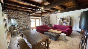 Un intérieur rustique avec un mur en pierre, des poutres en bois au plafond, une table à manger avec des chaises, un canapé rouge et un téléviseur.