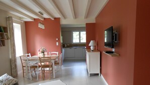 A living room with a white sofa, a dining table with chairs, a white coffee table, a TV on a stand, and a kitchen in the background.