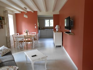 Una sala de estar con un sofá blanco, una mesa de comedor con sillas, una mesa baja blanca, un televisor sobre un mueble bajo y una cocina al fondo.