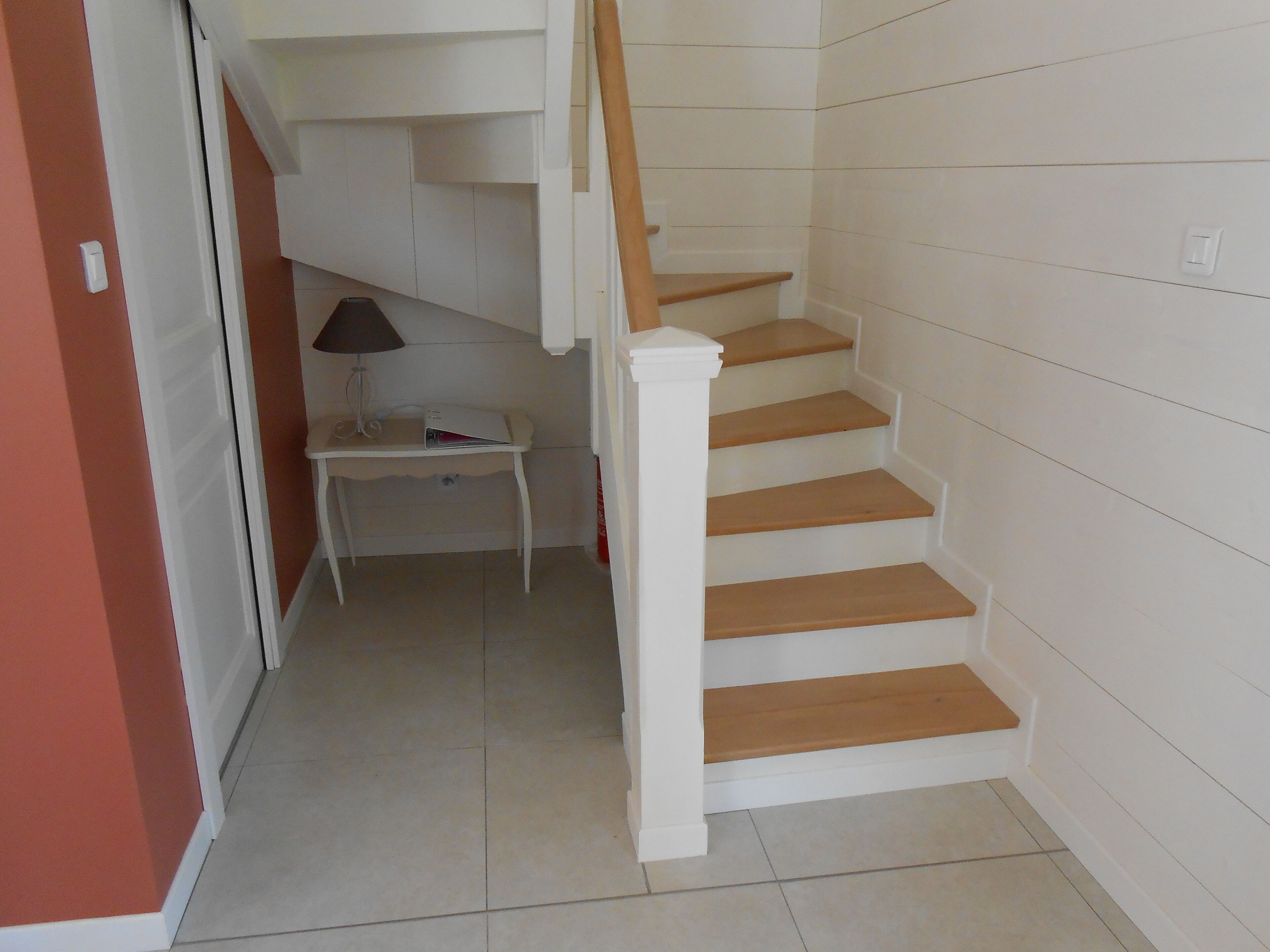 Eine Treppe mit Holzstufen und weißen Wänden führt zu einer Empore mit einem kleinen Tisch und einer Lampe.