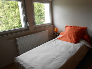 A bedroom with a bed, two windows, a radiator, and a nightstand with a lamp.