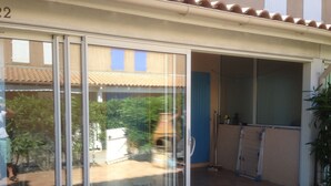 Une porte-fenêtre coulissante donne sur un espace patio doté d’un sol carrelé, d’une chaise en métal et d’une petite table.